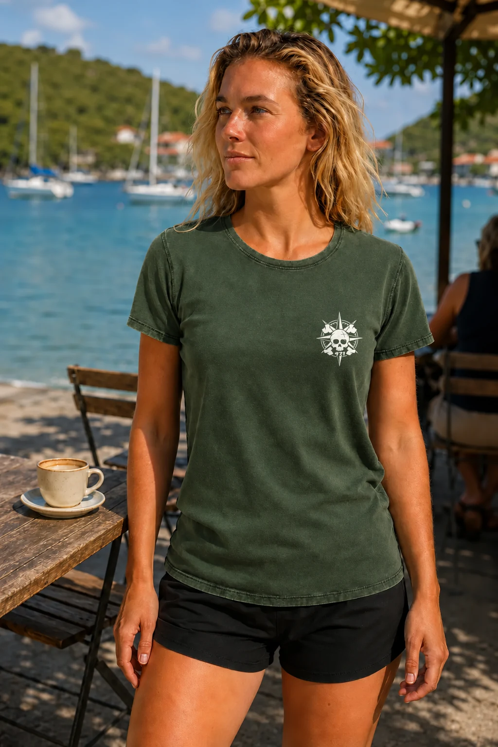 T-shirt vieilli femme vert militaire foncé délavé avec une boussole pirate 971 ornée d’un crâne imprimé en blanc, porté à la terrasse d’un café près de la baie des Saintes et de se