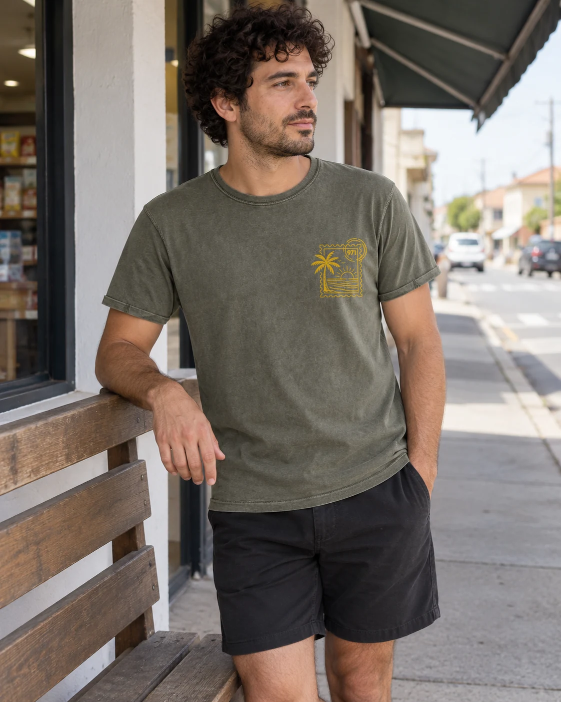 T-shirt vieilli homme vert militaire foncé délavé avec un timbre palmier 971 avec mer et soleil imprimé en jaune moutarde, porté par un homme sur le ponton d’un voilier aux Saintes